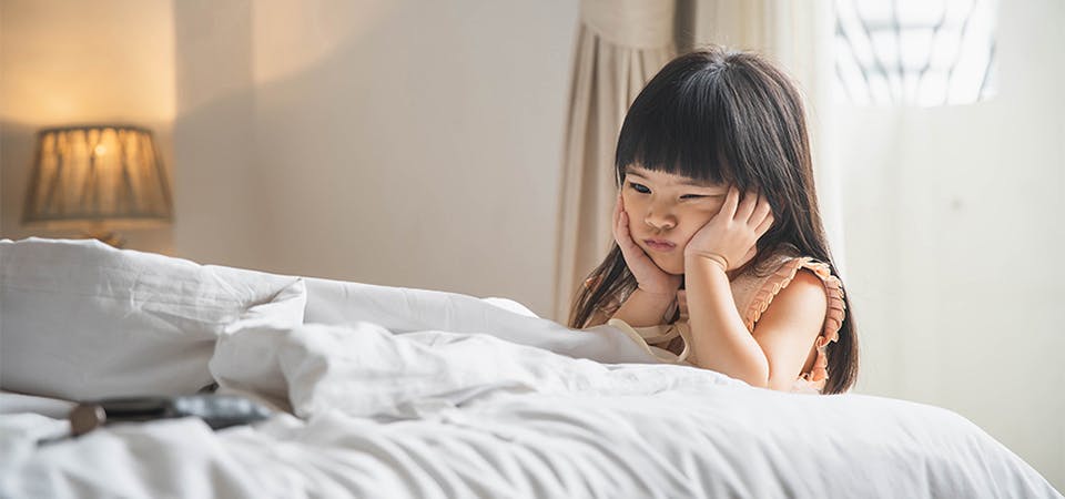 Little girl with hands on her cheeks looking mad and frustrated, kneeling down with elbows on bed inside of a room, alone.