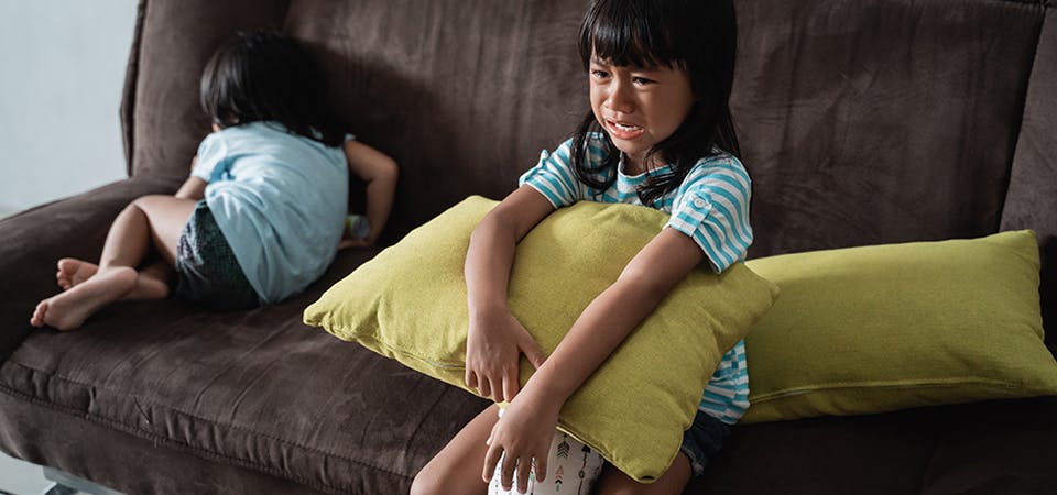 Two sisters on couch, one is crying holding a pillow and the other is curled up, turned away.