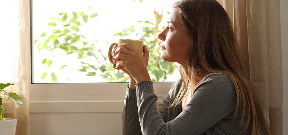 Young mom holding a cup of coffee, looking out the window in a deep state of thought and reflection.