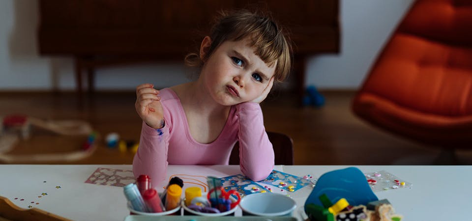 Little girl sitting at art table, resting her head in one hand and pouting, looking bored.