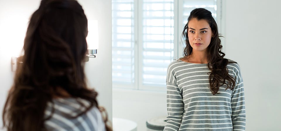 Brunette woman standing in front of mirror, looking at reflection thoughtfully