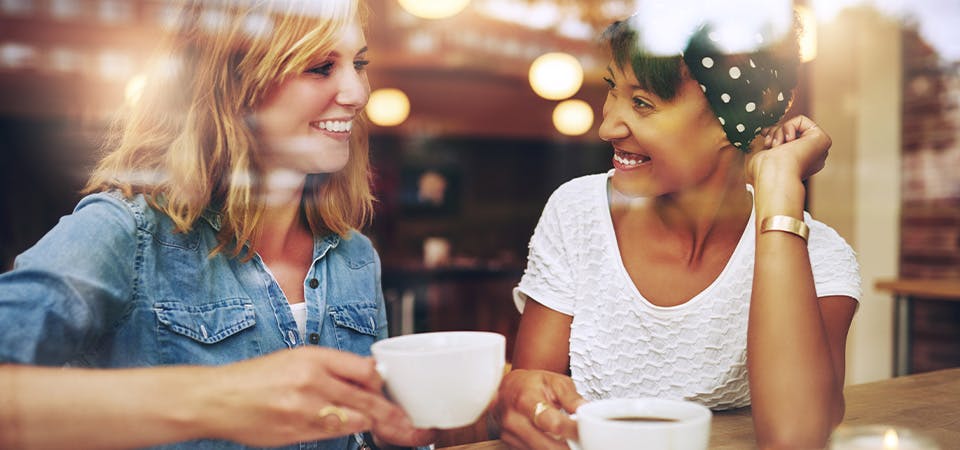 Two women smiling and drinking coffee together at coffee shop, seen through window