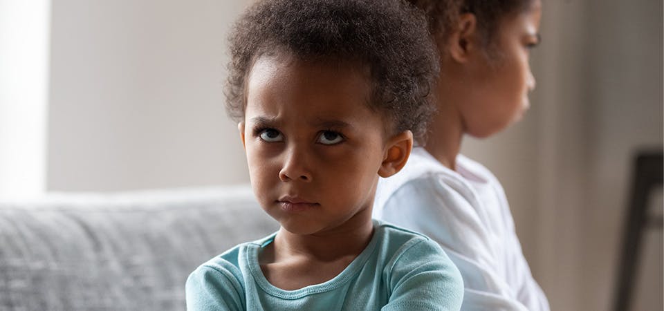 Two young african american girls sitting back-to-back on couch, looking angry/scowling