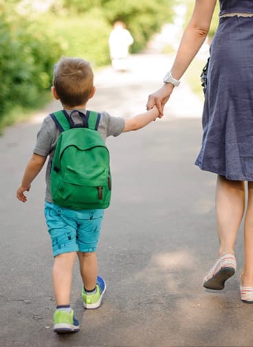 Young child with backpack on holding mother’s hand outside walking to school