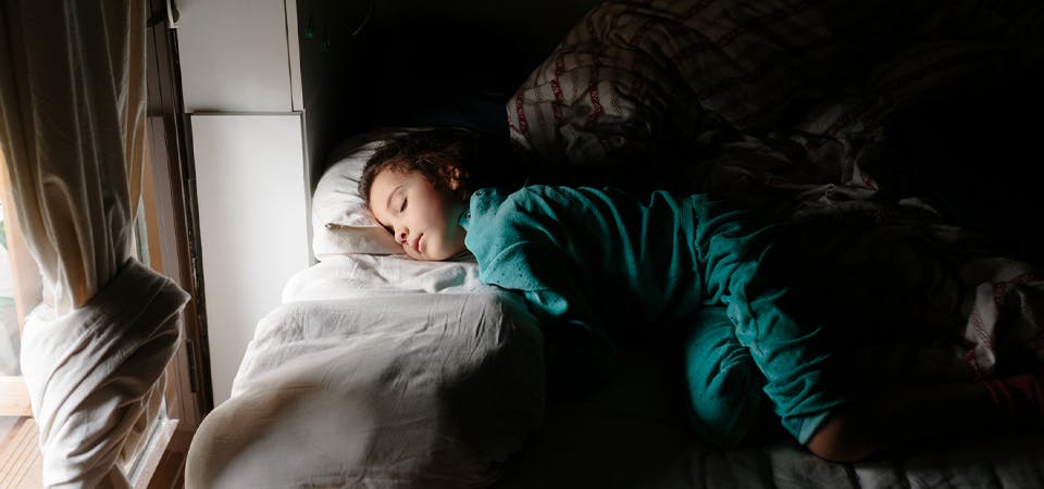 Kid sleeping on top of covers with light peeking through nearby window