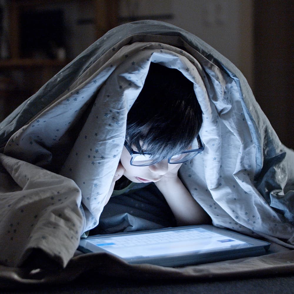 boy with black hair and glasses laying on his stomach covered in a blanket and looking at a tablet screen